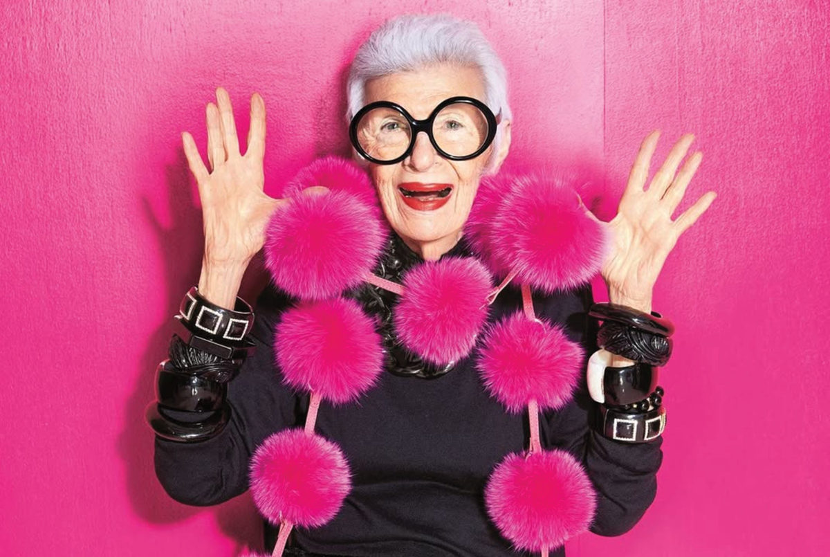 Iris Apfel s veľkými okuliarmi a ružovými brmbolcami na ružovom pozadí